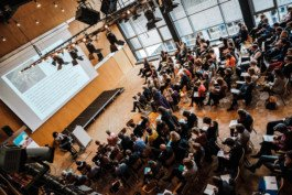 aufnahme von oben von eiem Konferenzraum mit vielen Menschen, die auf eine Rednerin schauen. Portrait von einem vollbärtigen Mann mit kurzen Haaren, der in die Kamera lächelt. Er trägt ein Shirt mit einem Fahrrad.