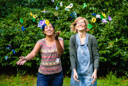 Zwei weiblich gelesen Personen werfen Buchstaben in die Luft Portrait von einem vollbärtigen Mann mit kurzen Haaren, der in die Kamera lächelt. Er trägt ein Shirt mit einem Fahrrad.