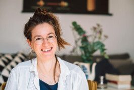 Portrait von Jana Kreisl eine weiblich gelesene Person, die eine Brille trägt und in die Kamera lächelt.