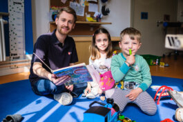 Zwei Kinder sitzen in einer Kita und schauen in die Kamera, eines trägt ein Diabetes Messgerät Im Hintergrund sitzt ein Malteser Begleiter mit einem Buch in der Hand Portrait von einem vollbärtigen Mann mit kurzen Haaren, der in die Kamera lächelt. Er trägt ein Shirt mit einem Fahrrad.