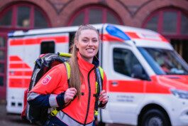 Eine weiblich gelesene Perosn mit roter Notarztkleidung udn einen Notarztrucksack lächelt vor einem Rettungswagen Portrait von einem vollbärtigen Mann mit kurzen Haaren, der in die Kamera lächelt. Er trägt ein Shirt mit einem Fahrrad.